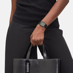 Eine Person in Schwarz, die ein ärmelloses Oberteil trägt und eine schwarze Handtasche in der Hand hält, trägt die elegante Liebeskind Berlin Armbanduhr - The Square Minimalist.