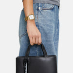 Eine Person in Jeans und einem gerippten grauen Oberteil trägt eine schwarze Handtasche und präsentiert die Liebeskind Berlin Armbanduhr - The Basic Staple an ihrem Handgelenk.