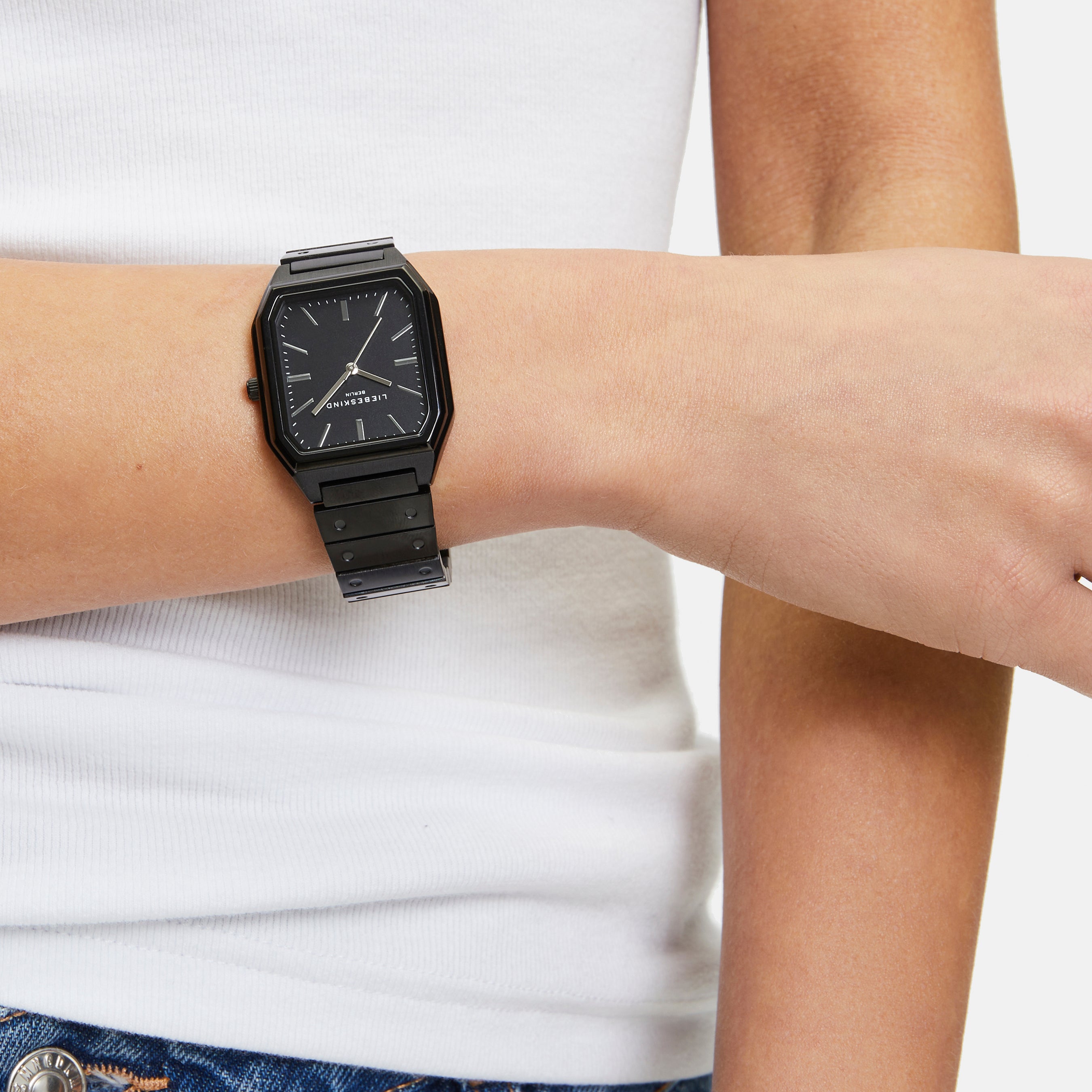 Eine Person trägt eine Liebeskind Berlin Armbanduhr - The Basic Staple, ein weißes Tanktop und eine blaue Jeans mit silbernen Knöpfen.