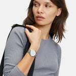 Eine Frau in einem grauen Rippenhemd trägt die Liebeskind Berlin Armbanduhr - The Round Staple und blickt mit einer Jacke über der Schulter zur Seite. Die silberne Uhr verleiht ihrem stilvollen Look ein modernes Design-Flair.