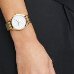 Die LIEBESKIND BERLIN Armbanduhr - The Round Staple überzeugt mit modernem Design, goldfarbenem Band und weißem Zifferblatt und setzt stilvolle Akzente zu schwarzer, strukturierter Kleidung.