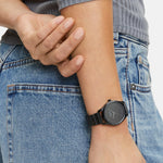 Eine Person trägt eine blaue Jeans, ein geripptes graues Top und die LIEBESKIND BERLIN Armbanduhr - The Round Staple von Liebeskind Berlin berührt ihren Arm.