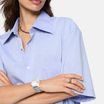 Frau in einem hellblauen Button-up-Hemd mit gekreuzten Armen, dazu die Liebeskind Berlin Armbanduhr - Round Vinyl Vision und silberne Ringe für einen Hauch von 70er-Jahre-Stil.