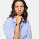 Eine Frau mit hellblauem Hemd trägt die LIEBESKIND BERLIN Armbanduhr - Round Vinyl Vision gold von Liebeskind Berlin mit verspiegeltem Ziffernblatt, während sie vor einem schlichten Hintergrund ihr Kinn auf ihre Hand stützt.