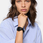 Eine Frau in einem hellblauen Hemd zeigt ihre Liebeskind Berlin Armbanduhr - Round Vinyl Vision, die Hand am Kinn und mit neutralem Gesichtsausdruck.