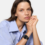 Eine Frau in einem hellblauen Hemd mit gefalteten Händen trägt die Liebeskind Berlin Armbanduhr - Round Vinyl Vision und schaut in die Kamera.