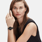 Eine Frau mit hellbraunen Haaren, die ein ärmelloses schwarzes Oberteil und die Liebeskind Berlin LIEBESKIND BERLIN Armbanduhr - Keep Loving trägt, schaut in die Kamera.