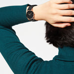 Eine Person im blaugrünen Pullover mit Hand auf dem Kopf trägt die LIEBESKIND BERLIN Armbanduhr - The Modern Must-Have von Liebeskind Berlin: eine schwarze Armbanduhr mit goldenem Gehäuse, schwarzem Zifferblatt und stylischem Echtleder-Armband.