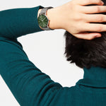 Eine Person im türkisen Sweater präsentiert am Handgelenk lässig nahe am Kopf die LIEBESKIND BERLIN Armbanduhr - Color Up mit grünem Zifferblatt, Edelstahl-Meshband und 5 ATM Wasserdichte der Marke Liebeskind Berlin.