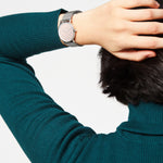 Eine Person in einem blaugrünen Pullover berührt ihren Kopf und trägt die LIEBESKIND BERLIN Armbanduhr - Color Up von Liebeskind Berlin, die sich durch minimalistisches skandinavisches Design und ein elegantes silbernes Netzband auszeichnet.