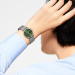 Eine Person trägt die LIEBESKIND BERLIN Armbanduhr - Color Up von Liebeskind Berlin mit grünem Zifferblatt, silbernem Mesh-Band, poliertem Edelstahl und minimalistischem skandinavischem Design, während sie ihre Hand an den Kopf hält.