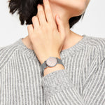 Eine Person in einem grau gestreiften Pullover berührt ihren Hals und trägt die LIEBESKIND BERLIN Armbanduhr - Color Up von Liebeskind Berlin, eine elegante Edelstahluhr mit Netzband und minimalistischem skandinavischem Design.