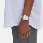 A person in a light blue shirt and gray pants wears the LIEBESKIND BERLIN Armbanduhr – The Square Minimalist on their left wrist, arm relaxed at their side, against a plain background.