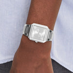 A close-up of a wrist wearing the LIEBESKIND BERLIN Armbanduhr – The Square Minimalist, featuring a silver square face and metal band, against a light blue shirt and dark pants.