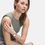 Eine Frau mit langen Haaren in einem ärmellosen Top und einer grauen Hose, die Schmuck und eine stilvolle Liebeskind Berlin Armbanduhr - The Classy with Edelstahl-Armband trägt, sitzt und schaut in die Kamera.