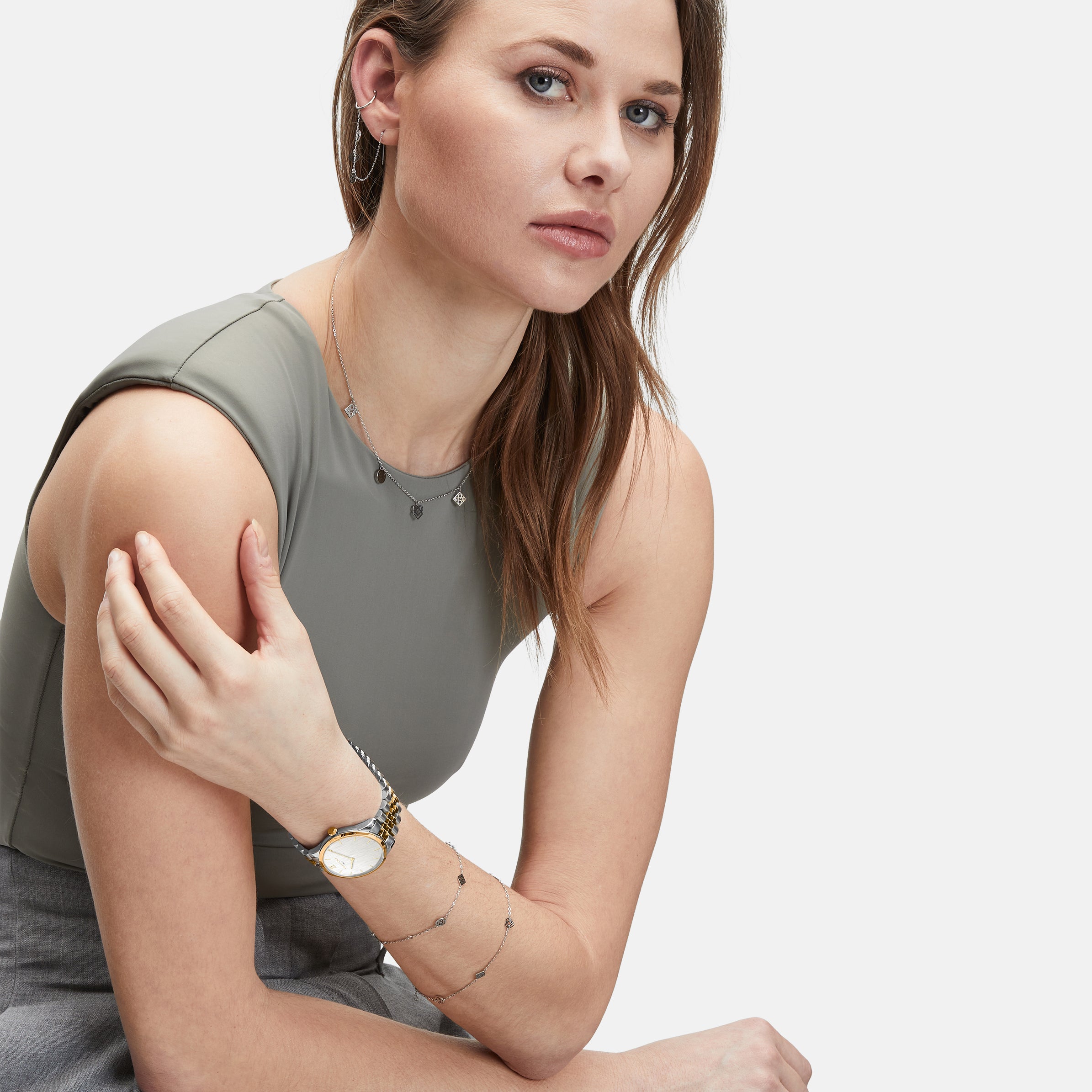 Frau mit hellbraunem Haar, in einem ärmellosen grauen Oberteil und einer Hose, sitzend und mit der LIEBESKIND BERLIN Armbanduhr - The Classy von Liebeskind Berlin sowie elegantem Schmuck.