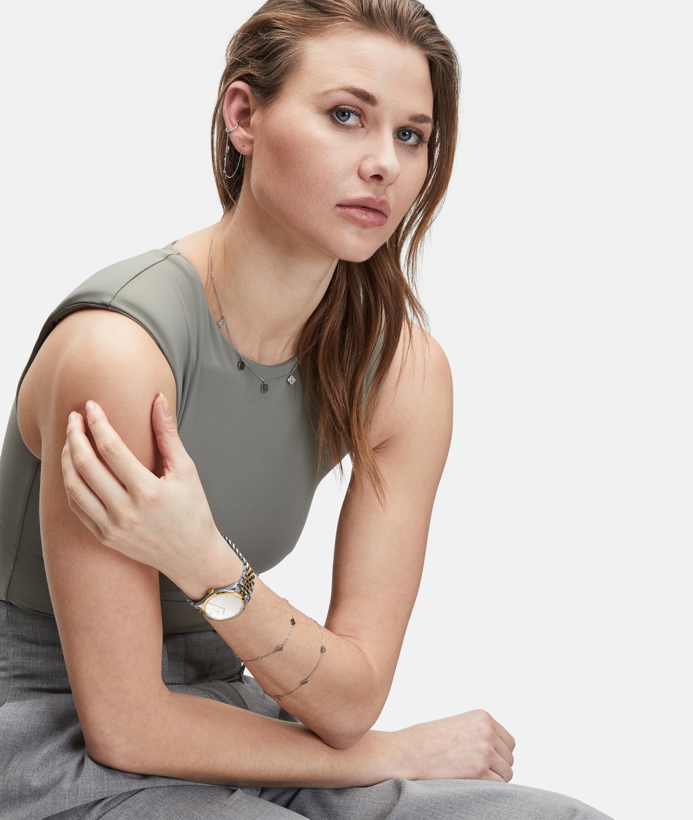 Frau mit hellbraunem Haar, in einem ärmellosen grauen Oberteil und einer Hose, sitzend und mit der LIEBESKIND BERLIN Armbanduhr - The Classy von Liebeskind Berlin sowie elegantem Schmuck.
