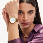 Eine Frau in einem lila Hemd modelliert die Liebeskind Berlin Armbanduhr - The Multi-Function mit goldenem Gehäuse und braunem Armband, während sie ihren Arm an ihr Gesicht hält.