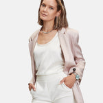 Eine Frau in einem hellrosa Blazer und weißem Outfit steht selbstbewusst mit den Händen in den Taschen und präsentiert die LIEBESKIND BERLIN Armbanduhr - Modern Chronograph von Liebeskind Berlin an ihrem Handgelenk.