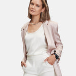 Eine Frau in einem hellrosa Blazer, einem weißen Oberteil und einer weißen Hose posiert mit den Händen in den Taschen, gestylt mit der Liebeskind Berlin Modern Chronograph Armbanduhr vor einem weißen Hintergrund.