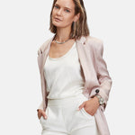 Frau in hellrosa Blazer und weißem Outfit, Hände in den Taschen, blickt selbstbewusst in die Kamera, während sie die stilvolle LIEBESKIND BERLIN Armbanduhr - Modern Chronograph von Liebeskind Berlin mit einem eleganten Edelstahlarmband trägt.