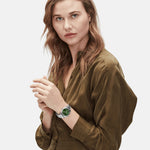 Eine Frau in einem olivgrünen Kleid trägt die LIEBESKIND BERLIN Armbanduhr - The Colorful Multifunction von Liebeskind Berlin mit grünem Zifferblatt und elegantem Edelstahlarmband vor einem weißen Hintergrund.