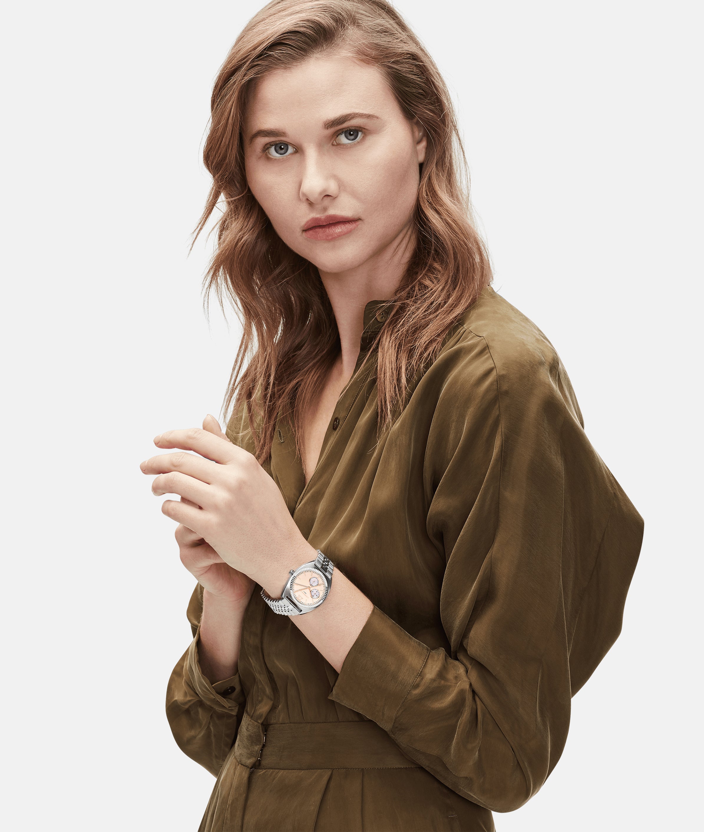Frau in einem olivgrünen Kleid, die die LIEBESKIND BERLIN Armbanduhr - The Colorful Multifunction von Liebeskind Berlin trägt, posiert vor einem weißen Hintergrund und schaut in die Kamera.