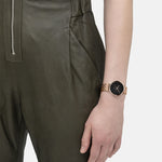 Eine Person in einem olivgrünen Overall trägt am linken Handgelenk eine Liebeskind Berlin Armbanduhr - The Colorful Must-Have, die eine goldene Armbanduhr mit schwarzem Zifferblatt zeigt.