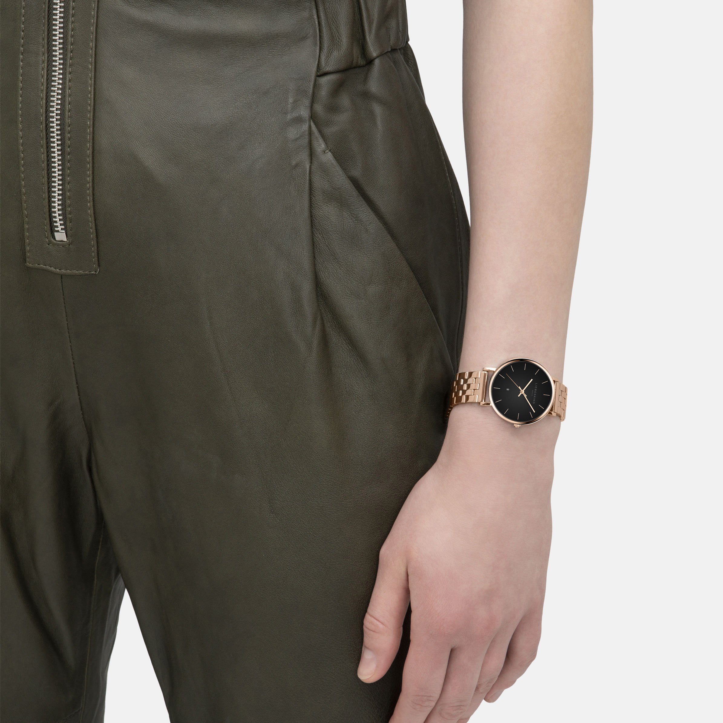 Eine Person in einem olivgrünen Overall trägt am linken Handgelenk eine Liebeskind Berlin Armbanduhr - The Colorful Must-Have, die eine goldene Armbanduhr mit schwarzem Zifferblatt zeigt.