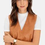 Eine Frau in brauner Weste und weißem Oberteil präsentiert eine Liebeskind Berlin Armbanduhr - The Classic Square in Gold an ihrem linken Handgelenk.