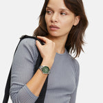 Frau in einem grauen gerippten Oberteil, die die LIEBESKIND BERLIN Armbanduhr - The Round Staple von Liebeskind Berlin trägt, ein schwarzes Band über der Schulter hält und zur Seite schaut.