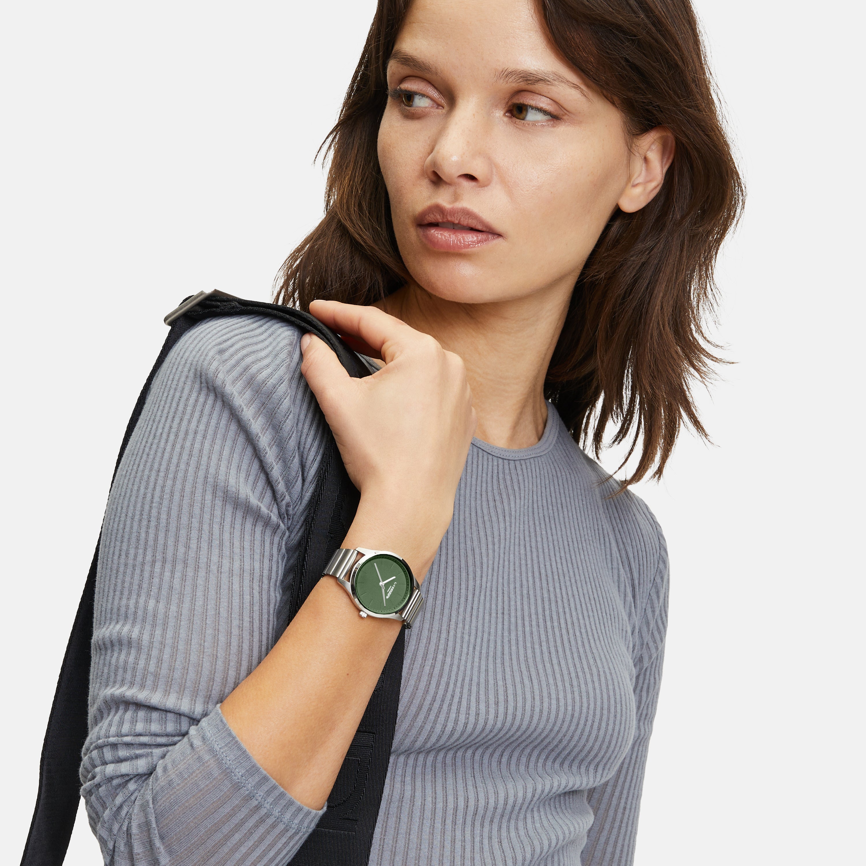 Frau in einem grauen gerippten Oberteil, die die LIEBESKIND BERLIN Armbanduhr - The Round Staple von Liebeskind Berlin trägt, ein schwarzes Band über der Schulter hält und zur Seite schaut.
