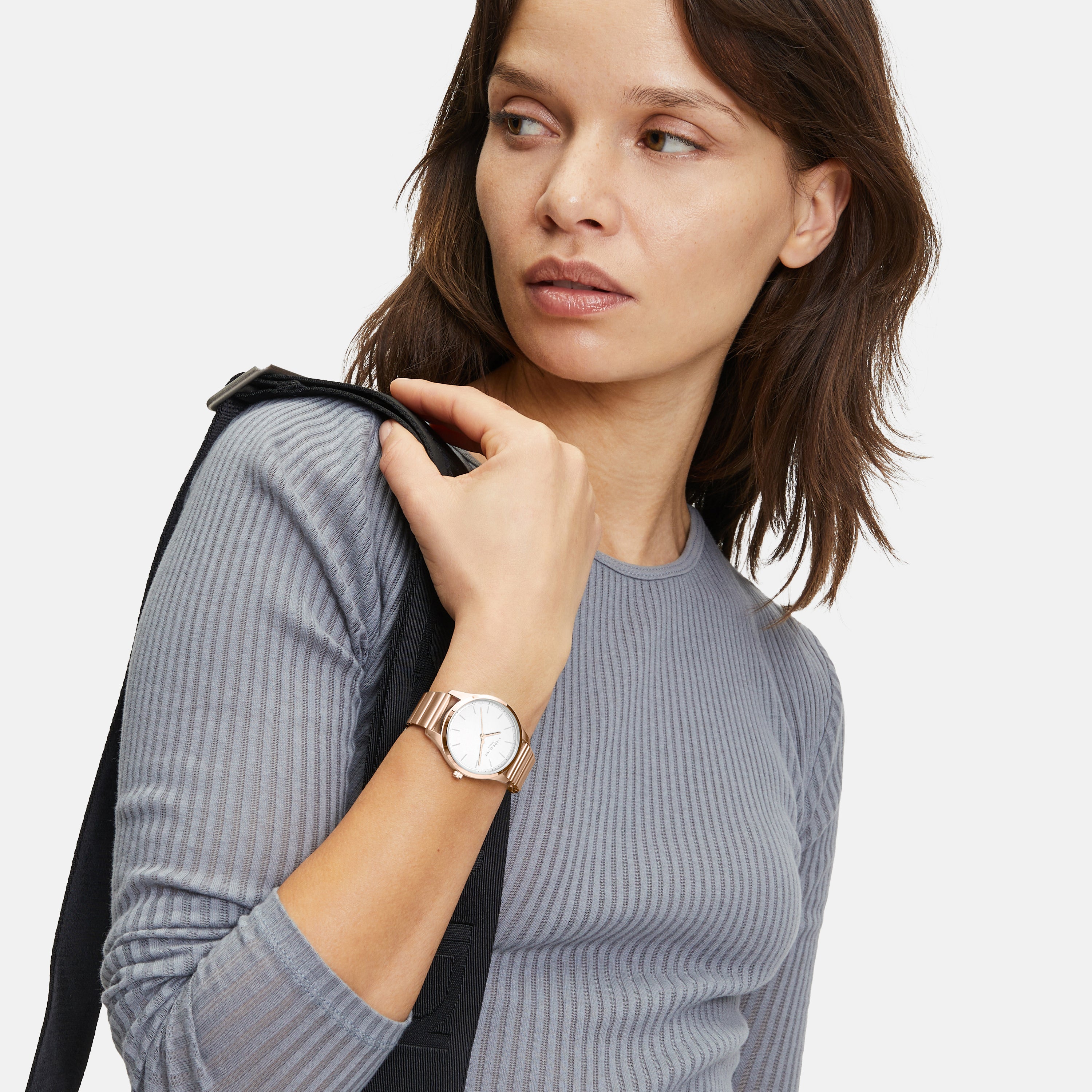 Frau im grauen Rippenhemd trägt die LIEBESKIND BERLIN Armbanduhr - The Round Staple von Liebeskind Berlin und hält eine schwarze Jacke über der Schulter.