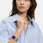 Frau in hellblauem Hemd trägt die Liebeskind Berlin Armbanduhr - The Modern Staple, eine elegante Quarzuhr mit blauem, quadratischem Zifferblatt, mit Blick zur Seite.