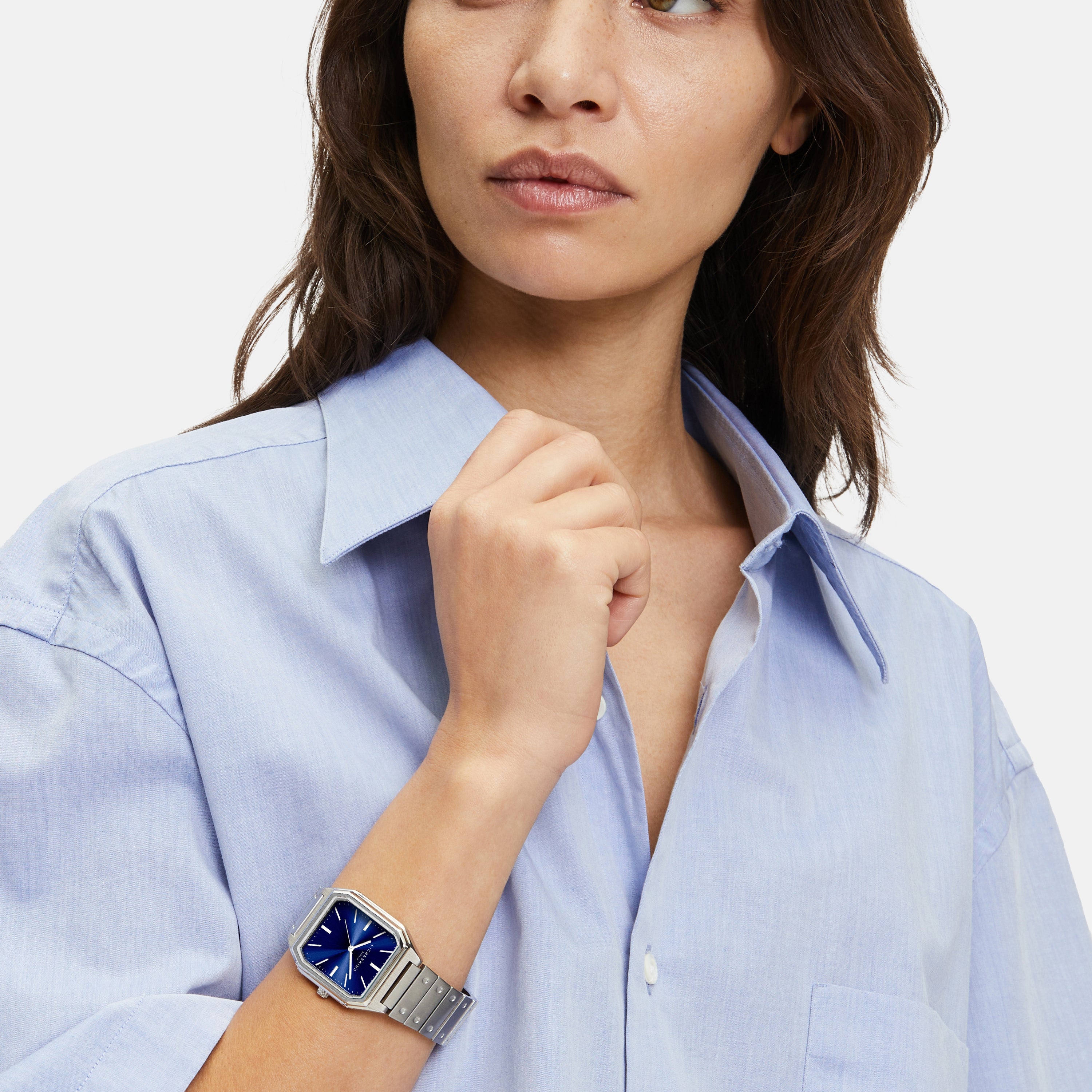 Frau in hellblauem Hemd trägt die Liebeskind Berlin Armbanduhr - The Modern Staple, eine elegante Quarzuhr mit blauem, quadratischem Zifferblatt, mit Blick zur Seite.