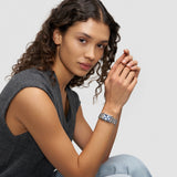 Frau mit lockigem Haar, die ein ärmelloses graues Top und eine Liebeskind Berlin Armbanduhr - Small Retro trägt, sitzt mit verschränkten Händen.