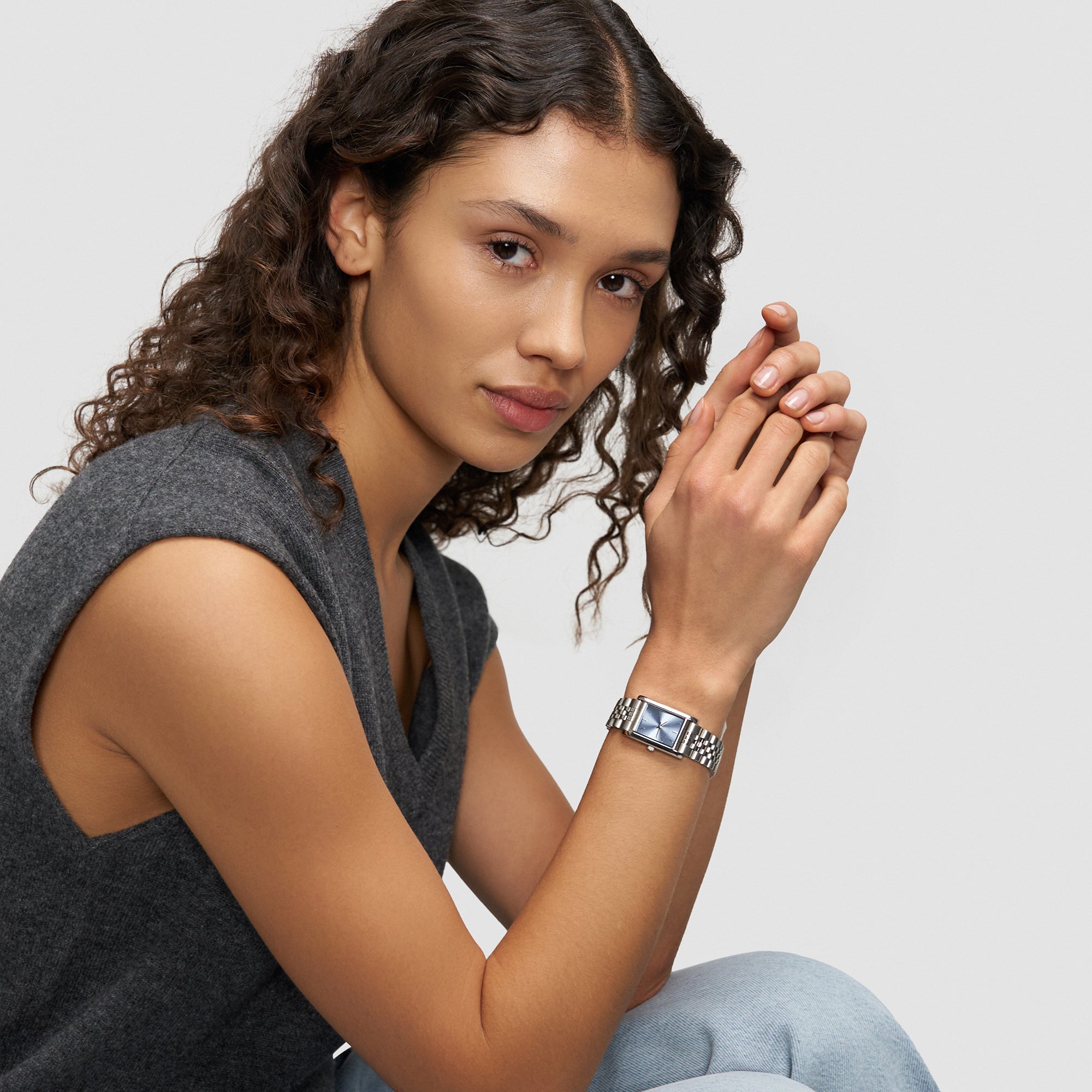 Frau mit lockigem Haar, die ein ärmelloses graues Top und eine Liebeskind Berlin Armbanduhr - Small Retro trägt, sitzt mit verschränkten Händen.