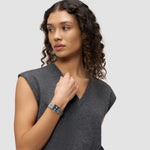 Frau mit lockigem Haar trägt ein graues ärmelloses Oberteil und die LIEBESKIND BERLIN Armbanduhr - Small Retro von Liebeskind Berlin, mit Blick zur Seite.