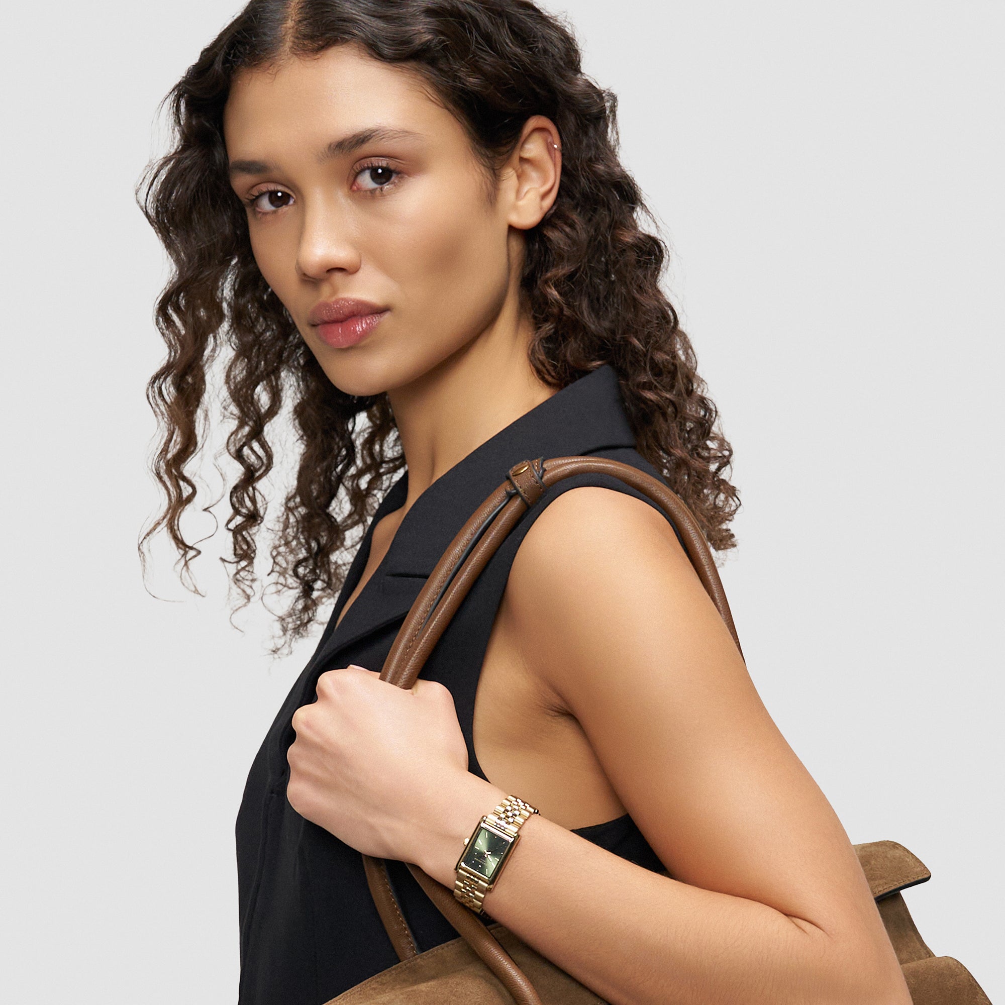 Frau mit lockigem Haar trägt ein schwarzes ärmelloses Oberteil, hat eine braune Tasche dabei und zeigt die LIEBESKIND BERLIN Armbanduhr - Small Retro von Liebeskind Berlin, die ihren schicken Stil perfekt unterstreicht.