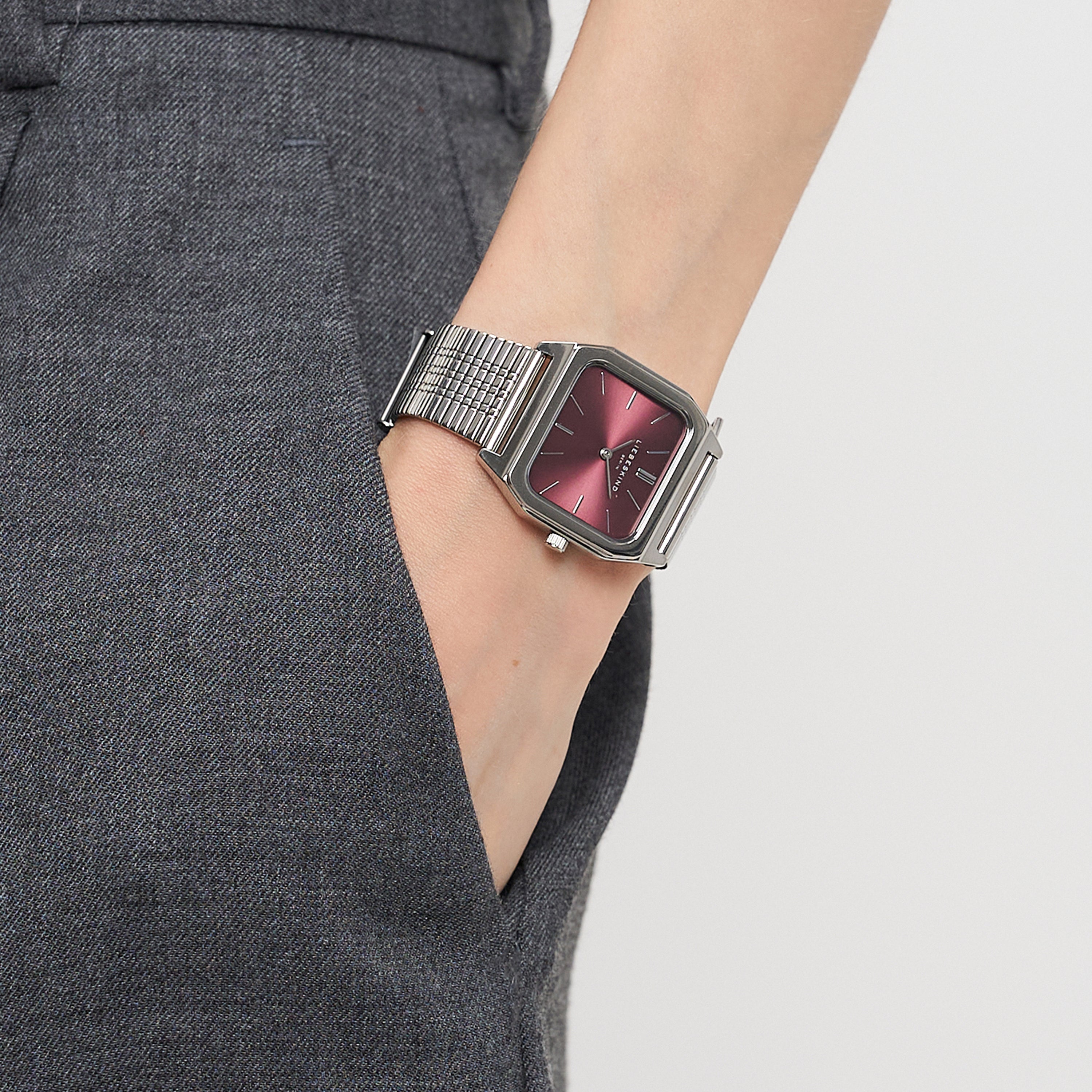 Person trägt eine graue Hose mit einer Liebeskind Berlin Armbanduhr - Vintage Classic mit rotem Zifferblatt und Retro-Stil. Das silberne Edelstahlmeshband verleiht dieser Vintage-inspirierten Uhr von Liebeskind Berlin einen modernen Touch.