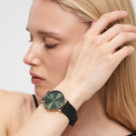 Eine Frau mit langen blonden Haaren trägt die LIEBESKIND BERLIN Armbanduhr - The Modern Must-Have von Liebeskind Berlin, eine schwarze Armbanduhr mit grünem Zifferblatt und poliertem Edelstahl, während sie ihren Kopf berührt.