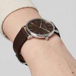 KI generierte Detailaufnahme einer LIEBESKIND BERLIN Damen Armbanduhr mit braunem Zifferblatt, silberfarbenem Edelstahlgehäuse und dunkelbraunem Lederarmband am Handgelenk getragen
