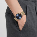 Eine Person trägt die LIEBESKIND BERLIN Armbanduhr - The Colorful Minimalist von Liebeskind Berlin, die ein minimalistisches Design, ein goldenes Edelstahl-Meshband, ein dunkelblaues Gesicht und ihre Hand in einer grauen Hosentasche hat.