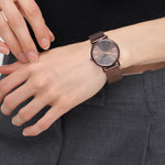 Eine Person in grauer Hose rückt ihre LIEBESKIND BERLIN Armbanduhr - The Everyday Must-Have, im minimalistischen skandinavischen Design mit braunem Meshband, am linken Handgelenk zurecht.