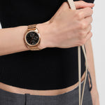 Eine Person, die die LIEBESKIND BERLIN Armbanduhr - The Multifunction von Liebeskind Berlin mit Edelstahlarmband trägt und ein beiges Armband hält, bekleidet mit einem schwarzen Oberteil und einer grauen Hose.