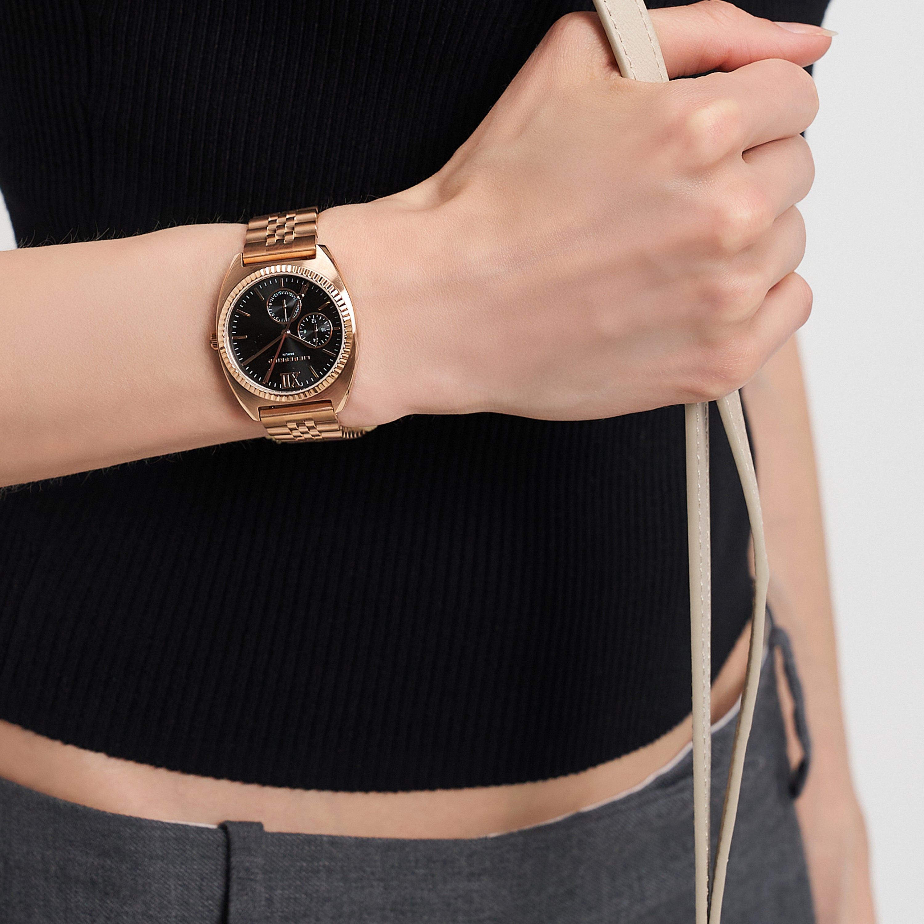 Eine Person, die die LIEBESKIND BERLIN Armbanduhr - The Multifunction von Liebeskind Berlin mit Edelstahlarmband trägt und ein beiges Armband hält, bekleidet mit einem schwarzen Oberteil und einer grauen Hose.