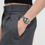 Person in grauer Hose trägt eine LIEBESKIND BERLIN Armbanduhr - The Classic Must-Have mit grünem Zifferblatt und Edelstahlband von Liebeskind Berlin, Hand in der Tasche.