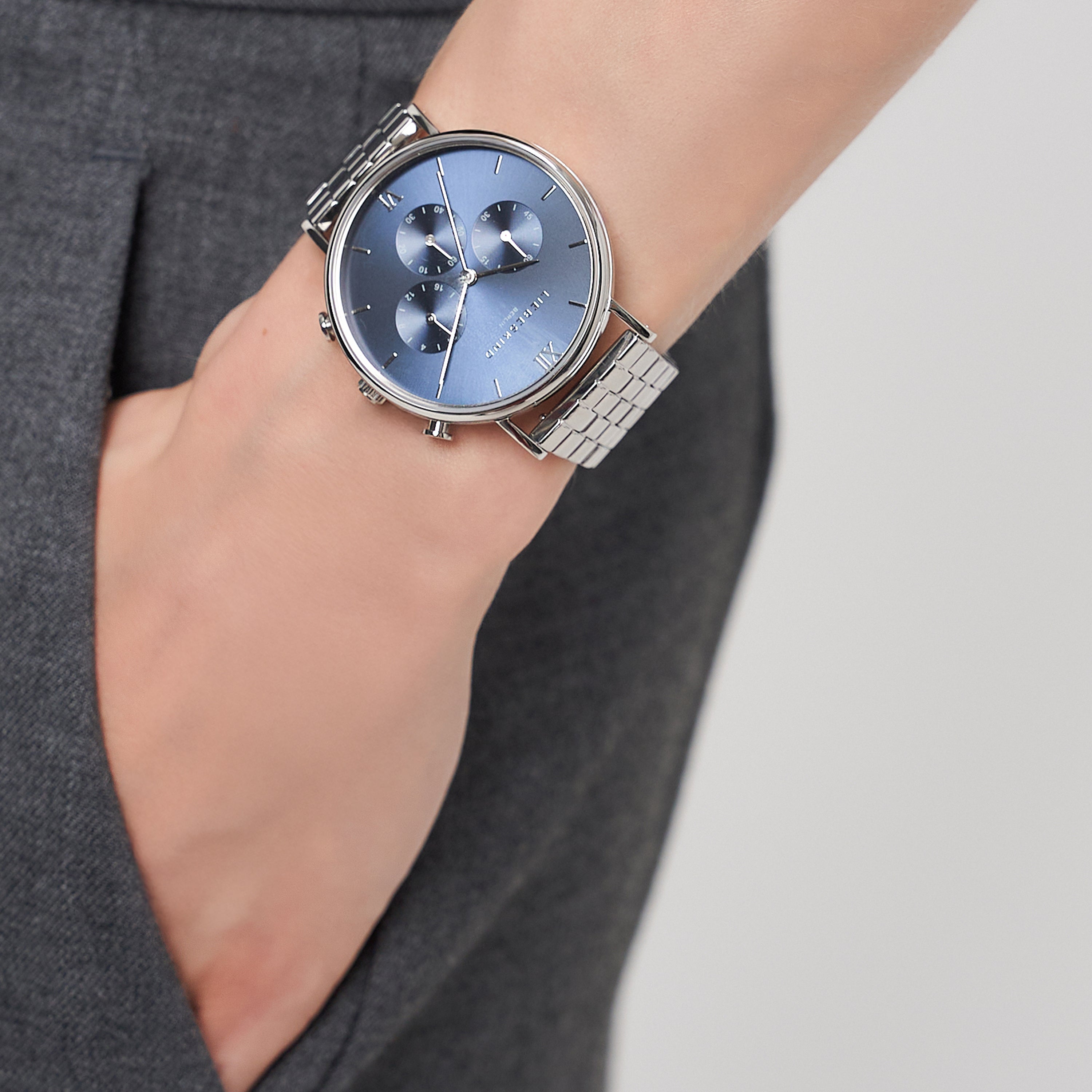 Eine Person trägt eine LIEBESKIND BERLIN Armbanduhr - Modern Chronograph von Liebeskind Berlin mit silbernem Gehäuse, blauem Zifferblatt und Edelstahlarmband, Hand in der Tasche.