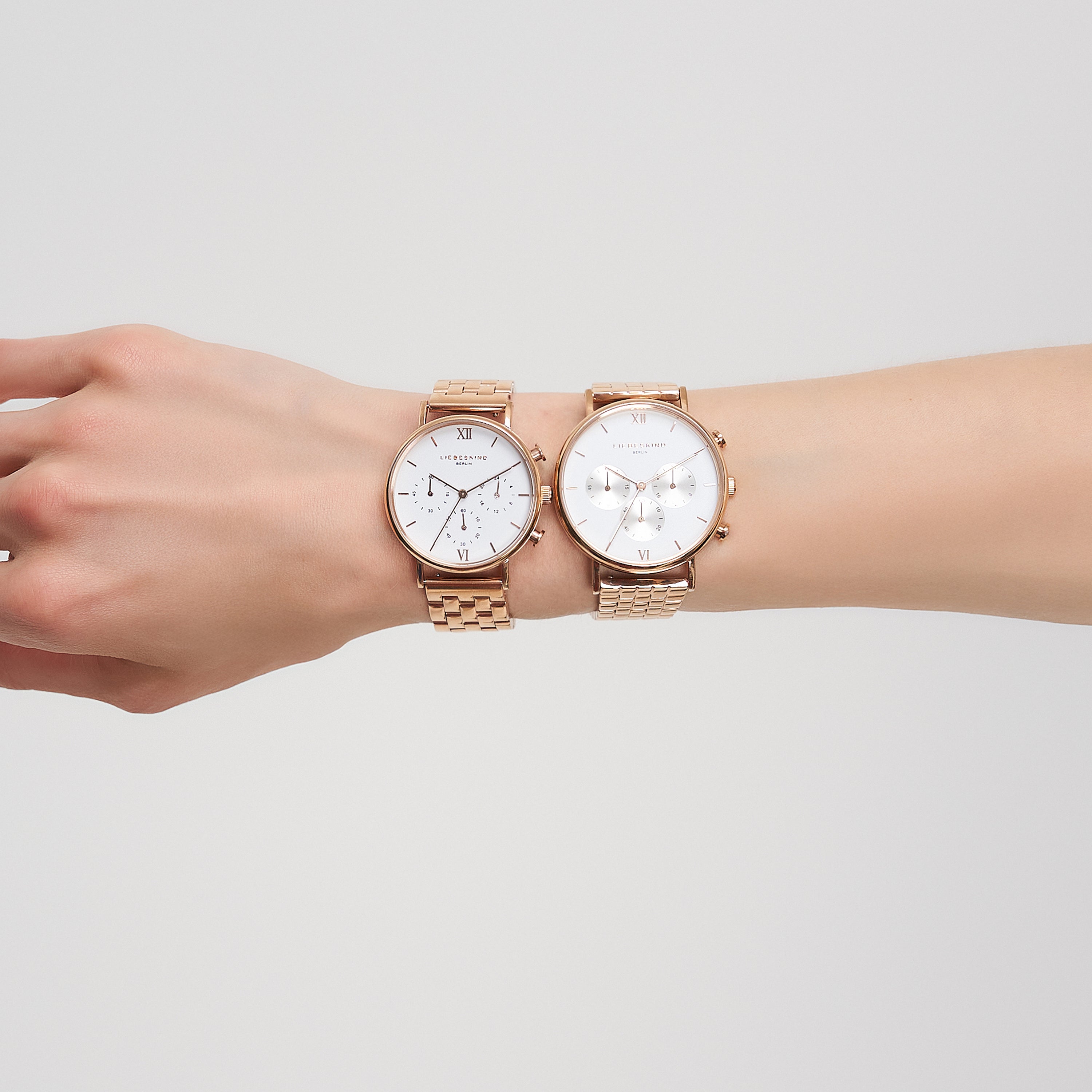 Eine Hand trägt zwei goldene Armbanduhren nebeneinander, darunter die stilvolle Liebeskind Berlin Armbanduhr - Small Modern Chronograph, vor einem hellen, neutralen Hintergrund.