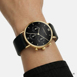 Ki generiertes Bild einer LIEBESKIND BERLIN Multifunktionsuhr mit goldfarbenem Edelstahlgehäuse, schwarzem Zifferblatt und schwarzem Lederarmband, getragen am Handgelenk.